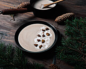 From above of black bowls with cream soup of chestnuts placed on wooden table with green branches of fir with cones
