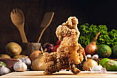 Tasty crispy fried chicken served on wooden cutting board on table with assorted vegetables in kitchen