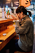 Junge asiatische Frau im Pullover isst Ramen mit Löffel an einem Holztresen in einem Cafe