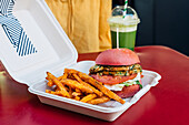 Leckerer Burger mit Brötchen und frischen Gemüsescheiben neben frittierten Kartoffelstäbchen in einer Box zum Mitnehmen auf dem Tisch im Restaurant