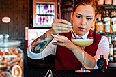 Professionelle junge Barkeeperin, die Alkohol aus einer Flasche in Form eines Totenkopfes mit einem Tropfer in ein Glas gibt, während sie einen sauren Cocktail in einer Bar zubereitet