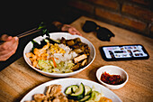 Frau in Freizeitkleidung mit Smartphone und Sonnenbrille isst leckeres taiwanesisches Bento mit Stäbchen in einem asiatischen Cafe