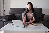 Fokussierte asiatische Frau schaut auf den Bildschirm eines Netbooks auf dem Tisch mit einem Frühstück bestehend aus Schokolade und Gebäck, während sie auf einer bequemen Couch sitzt.