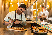 Male cook in uniform and mask for protecting from COVID 19 with grater adding cheese on flatbread pizza served on table with appetizers