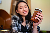 Young ethnic female with glass jar of delicious fig marmalade on table in house on blurred background