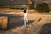 Back view of unrecognizable female throwing hat and running along dried field in summer evening in countryside