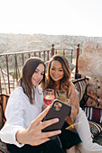 Hoher Winkel von ethnischen glücklichen Freundinnen, die auf Kissen sitzen, während sie ein Selbstporträt mit dem Handy am Tisch mit Sofortbildkamera und Cocktail auf der Terrasse in Kappadokien, Türkei, aufnehmen