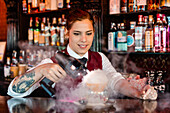 Skilled young female barkeeper using flavor bluster smoke gun while garnishing cocktail at bar counter