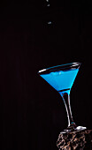 Drops of blue Lagoon cocktail pouring in crystal elegant glass placed on rough surface against black background
