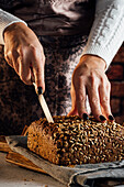 Anonyme Bäckerin mit Messer schneidet frisches Brot mit Sonnenblumenkernen auf einem Tisch in einer Backstube