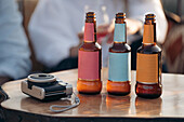 Crop anonymous friends sitting at table with glass bottles of beer near instant photo camera in bar