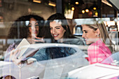Through glass of cheerful young multiracial female best friends in trendy outfits smiling and showing two fingers gesture while taking selfie on smartphone standing near window