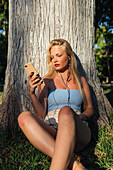 Unbeschwerte Frau, die mit ihrem Smartphone telefoniert und über Kopfhörer Musik hört, während sie im Sommer bei Sonnenuntergang unter einem Baum im Park sitzt und in die Kamera schaut