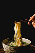 Hand of woman holding bamboo chopsticks with tasty wheat noodles from Chinese ramen meal plate