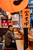 Asian woman in casual wear sitting at wooden counter while waiting for order in ramen bar