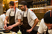 Männliche Köche in eleganter Uniform und mit Schutzmasken zur Verhinderung der Ausbreitung von COVID 19 bei der Zubereitung einer Mahlzeit in der Küche eines Restaurants (durch Glas)