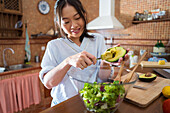 Glad Asian woman in casual clothes smiling and putting avocado cubes into glass bowl with lettuce while cooking vegan salad for lunch in sunlit kitchen