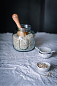 A clear glass jar with milled oats and a wooden scoop stands on a kitchen table covered with a linen cloth, flanked by smaller bowls with oat flakes