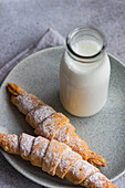 From above of two homemade jam-filled pastry cones with a dusting of powdered sugar, accompanied by a bottle of milk, on a ceramic plate over a grey napkin and countertop