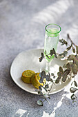 Delicate eucalyptus branches accentuate a slender glass of pistachio vodka and two textured pistachio cookies on a white plate atop a concrete surface