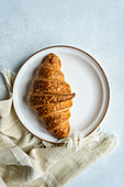 Draufsicht auf ein einzelnes Croissant auf einem runden Teller, das auf einem hessischen Tuch mit strukturiertem Hintergrund ruht und die Einfachheit eines Frühstücks mit Gebäck einfängt