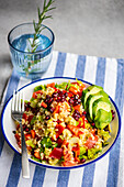 A colorful plate of bulgur salad with pomegranate seeds, fresh vegetables, and avocado slices, presented on a blue-striped linen, with a glass of water and rosemary sprig