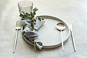 A modern and elegant table setting featuring a gray napkin, cutlery, and greenery on a stone charger