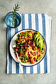 Top view of colorful plate of bulgur salad with pomegranate seeds, fresh vegetables, and avocado slices, presented on a blue-striped linen, with a glass of water and rosemary sprig
