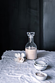 A serene composition featuring a glass bottle of homemade oat milk, a delicate flower accent, and scattered oats, all bathed in soft, natural light