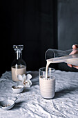 Anonymous hand pours creamy homemade oat milk from a clear jug into a glass, with a bottle and oats on a textured cloth