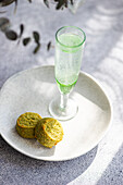 A flute glass filled with pistachio-flavored vodka stands beside two round pistachio cookies on a ceramic plate, all presented on a textured concrete table with soft natural lighting