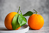 Two ripe oranges with vibrant green leaves, one in focus in the foreground on a concrete base with a blurred background