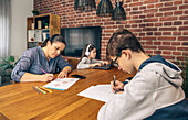 A family engages in various activities at the dining table: a woman colors, a girl uses a tablet with headphones, and a boy writes notes