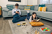 A teenage boy listens to music on his phone while his sister reads a book on the floor, surrounded by games and snacks in a cozy living room
