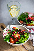 Top view of bowl of fresh vegetable salad with lettuce, arugula, radishes, cherry tomatoes, and pomegranate seeds on a wooden surface, beside a glass of lemon water