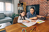 A woman assists a young girl with coloring while working from a laptop at a wooden dining table at home