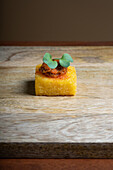 A gourmet polenta canapé topped with a rich tomato and onion stew and a sprig of fresh green garnish, elegantly presented on a wooden board.