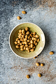 Top view of bowl of salty coated peanuts on a textured surface with some scattered around the bowl