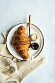 Top view of elegant breakfast plate with a croissant, jars of lemon ginger honey and cocoa hazelnut spread, accompanied by a knife on a woven cloth