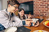 Geschwister lachen gemeinsam, während sie beim Familienfrühstück auf ein Smartphone schauen und einen gesunden Brotaufstrich auf den Tisch stellen