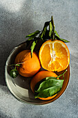 A ceramic plate holding fresh oranges, one sliced, with lush green leaves under natural light on a textured gray surface