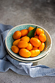 Top view of bowl of vibrant, fresh kumquats with a green leaf, placed on a textured surface with a blue napkin