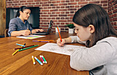 A focused child colors with crayons at a table while a woman works on a laptop in a home environment