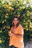 A cheerful young woman in a casual orange shirt picks fresh lemons from a lush tree in a sunlit home garden, embodying the joy of gardening.
