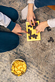Top view of two people playing a wooden puzzle game on the floor with a bowl of chips nearby, enjoying a family weekend at home