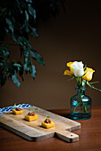 Polenta-Canapé mit Tomaten-Zwiebel-Eintopf, elegant präsentiert auf einem Holzbrett vor dem Hintergrund gelber Rosen in einer blauen Vase.