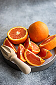 Ein Teller voller reifer, frisch geschnittener Orangen neben einer hölzernen Zitruspresse auf einem strukturierten grauen Hintergrund.