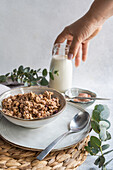 A hand pours milk into a glass beside a bowl of granola, with a spoon and eucalyptus leaves for decoration