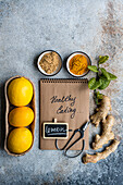 A notepad labeled healthy Cooking with a pen, fresh lemons, ginger, basil, and spices on a textured table