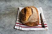 Top view of golden-brown rye sourdough bread on a white cloth with red stripes, next to a bread knife on a textured surface
